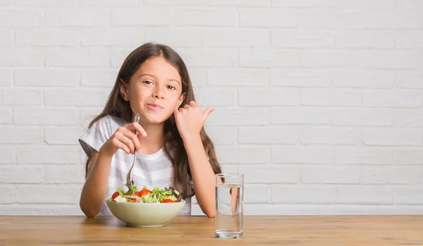 指していると幸せそうな顔を笑顔で側まで親指で示す健康的なサラダを食べるテーブルの上に座って若いヒスパニックの子供 — ストック写真