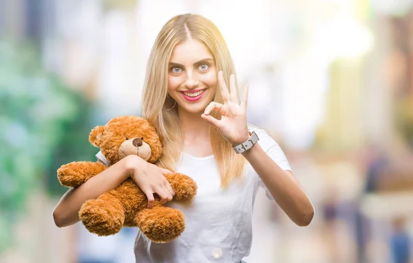 Young beautiful blonde woman holding teddy bear over isolated background doing ok sign with fingers, excellent symbol