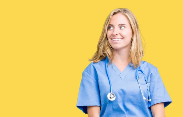 美丽的年轻医生妇女穿着医疗制服在孤立的背景微笑看一边 盯着思考 — 图库照片