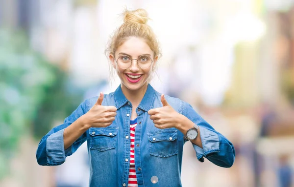 Young beautiful blonde woman wearing glasses over isolated background success sign doing positive gesture with hand, thumbs up smiling and happy. Looking at the camera with cheerful expression, winner gesture.