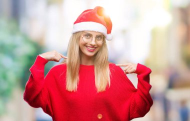 Yüzünde gülümseme ile kendinden emin görünüyorsun, kendini gururlu ve mutlu parmakları ile işaret izole arka plan üzerinde Noel şapka giyen genç güzel sarışın kadın.