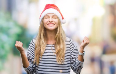 Genç güzel sarışın kadın izole arka plan kutlama üzerinde Noel şapka şaşırttı ve başarı için kaldırdı kollarını açarak hayrete ve gözleri açık. Kazanan kavramı.