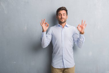 Yakışıklı genç iş adamı şık gömlek giymiş gri grunge duvarın üzerinden rahatla ve gülümseyen gözlerle kapalı parmak jestle meditasyon yapıyor. Yoga kavramı.