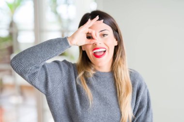 Genç güzel kadın giyiyor kış kazak evde el gülümseyen, ok jest yapıyor göz parmakları ile mutlu yüz üzerinden seyir.