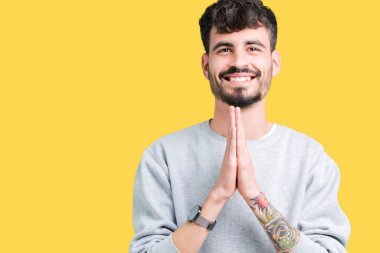 Ellerle birlikte AF emin gülümseyen için soran dua izole arka plan üzerinde sweatshirt giyen genç yakışıklı adam.