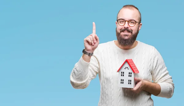 Young caucasian real state agent man holding house isolated background surprised with an idea or question pointing finger with happy face, number one