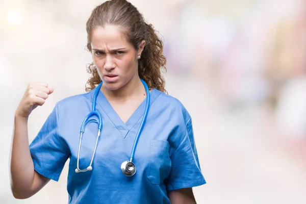 Young brunette doctor girl wearing nurse or surgeon uniform over isolated background angry and mad raising fist frustrated and furious while shouting with anger. Rage and aggressive concept.