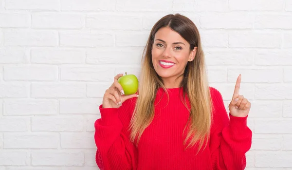 年轻的成年妇女在白色砖墙上吃新鲜的绿色苹果惊讶的想法或问题用快乐的脸指指点点 — 图库照片