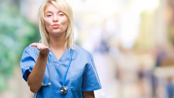 Young beautiful blonde doctor woman wearing medical uniform over isolated background looking at the camera blowing a kiss with hand on air being lovely and sexy. Love expression.