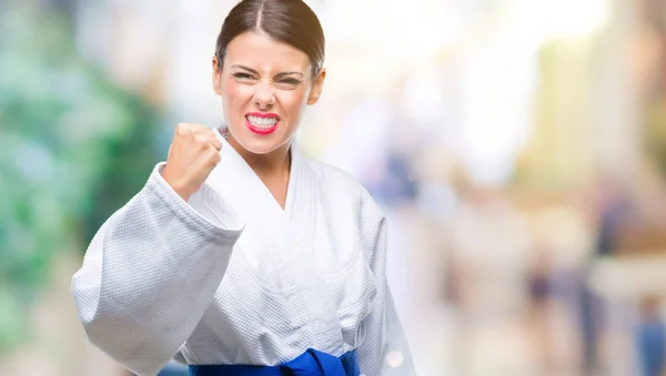 Young beautiful woman wearing karate kimono uniform over isolated background angry and mad raising fist frustrated and furious while shouting with anger. Rage and aggressive concept.