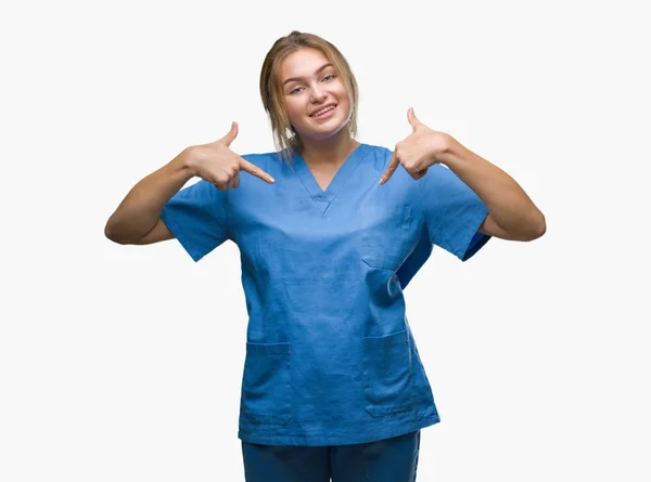 若い白人の看護師女性の顔に笑顔と自信を持って 誇りと幸せな指で自分を指して孤立の背景の上の外科医の制服を着て — ストック写真