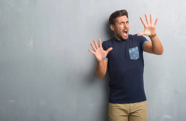 Handsome young man over grey grunge wall afraid and terrified with fear expression stop gesture with hands, shouting in shock. Panic concept.