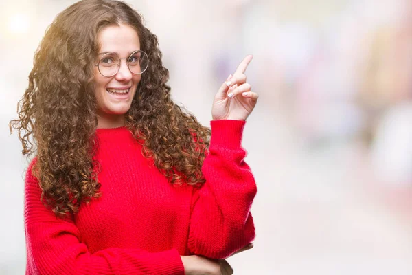 Kameraya bakarak yan el ve parmak ile işaret yüzünde kocaman bir gülümseme ile izole arka plan üzerinde gözlük ve kış kazak giyen güzel esmer kıvırcık saçlı Genç kız.