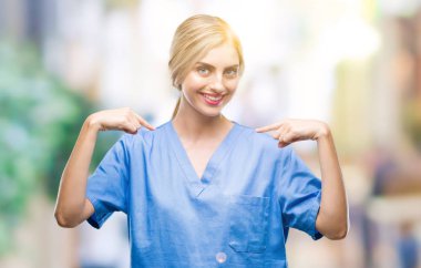 Genç güzel sarışın doktor cerrah hemşire kadın yüzünde gülümseme ile kendinden emin görünüyorsun, kendini gururlu ve mutlu parmakları ile işaret arka plan izole.