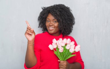 Gri grunge genç Afro-Amerikan kadın pembe çiçekler Buket sürpriz bir fikir ile tutarak duvar veya gülen yüz, bir numara ile işaret parmağı soru