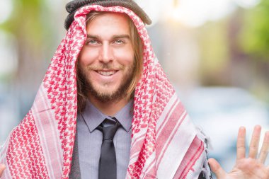Uzun saçlı keffiyeh gösterilen ve parmakları ile işaret izole arka plan üzerinde giyen genç yakışıklı Arap adam kendinden emin ve mutlu gülümseyerek süre on numara.