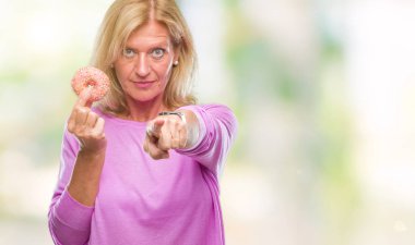 Orta yaş sarışın kadın üzerinde pembe çörek yeme kamera ve size parmak ile işaret arka plan izole, el işareti, pozitif ve kendinden emin bir jest ön