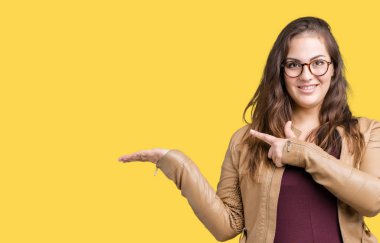 Hayret ve el ile sunulması ve parmak ile işaret ederken kameraya gülümseyen izole arka plan üzerinde elbise ve deri ceket giyen güzel artı boyutu genç kadın.