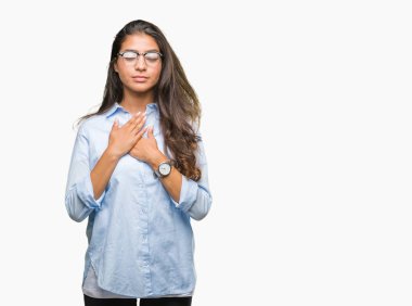 Eller genç güzel Arap kadın gözlük ile gülümseyen izole arka plan üzerinde kapalı gözler ve yüzünde minnettar jest ile göğüs üzerinde. Sağlık kavramı.