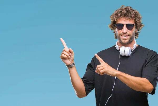 英俊的西班牙男子听音乐戴耳机在孤立的背景微笑着 看着相机指向镜头 用两只手和手指指着一边 — 图库照片