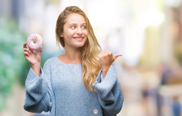 指していると幸せそうな顔を笑顔で側まで親指で示す分離の背景に甘いドーナツを食べる若い美しいブロンドの女性 — ストック写真