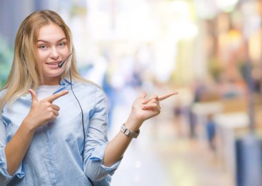 Gülümseyen ve iki elleri ve parmakları ile tarafına işaret kamera bakarak izole arka plan üzerinde çağrı merkezi kulaklık giyen genç beyaz iş kadın.