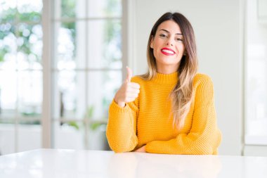 Young beautiful woman wearing winter sweater at home doing happy thumbs up gesture with hand. Approving expression looking at the camera showing success. clipart