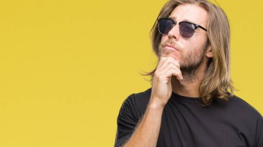Genç yakışıklı adam uzun saçlı çene düşünme hakkında soru, dalgın ifade üzerine el ile izole arka plan üzerinde güneş gözlüğü takıyor. Düşünceli yüzüyle gülümsüyor. Şüphe kavramı.