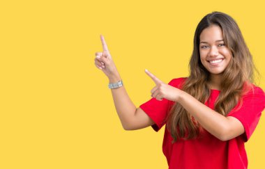 Gülümseyen ve iki elleri ve parmakları ile tarafına işaret kamera bakarak izole arka plan üzerinde kırmızı t-shirt giyen genç güzel esmer kadın.