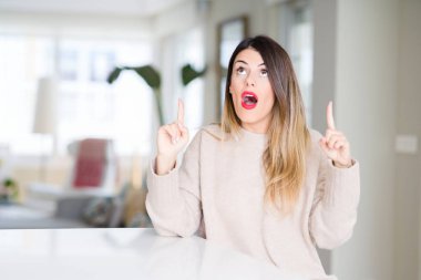 Evde kış kazak giyen genç güzel kadın hayrete ve yukarıya bakıyor ve parmakları ile işaret ve silah kaldırdı şaşırttı.