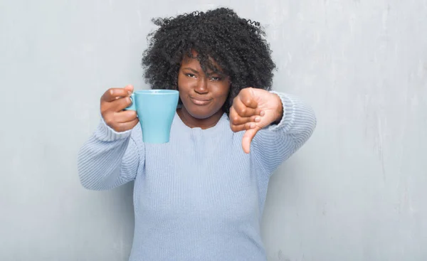 Young african american woman over grey grunge wall drinking a cup of coffee with angry face, negative sign showing dislike with thumbs down, rejection concept