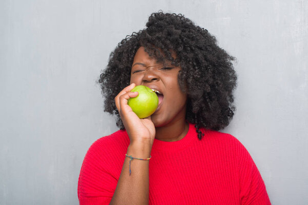 Young african american woman over grey grunge wall eating green apple with a confident expression on smart face thinking serious