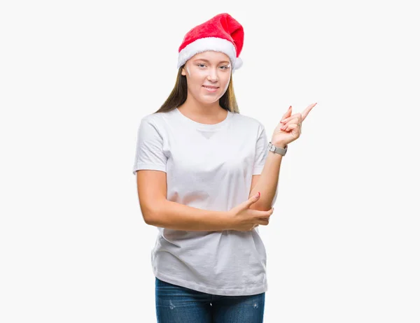若い美しい白人女性の顔に大きな笑みを浮かべて隔離された背景にクリスマスの帽子を着てカメラを見て側の手と指で指す — ストック写真