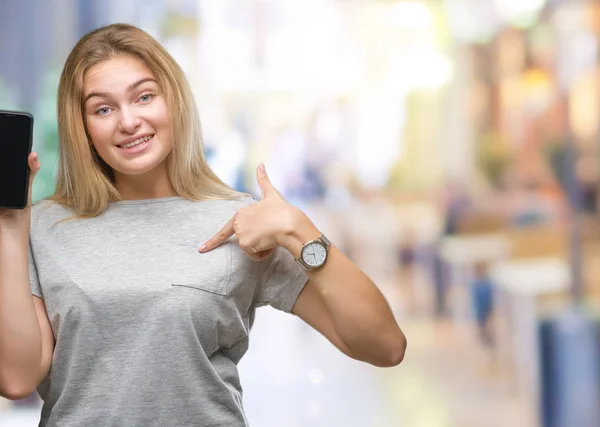 Young caucasian woman showing screen of smartphone over isolated background with surprise face pointing finger to himself