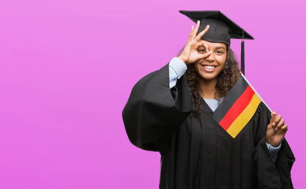 卒業のドイツの国旗を保持して指を通して見る目に手で サインをして笑っている幸せそうな顔で制服を着ている若いヒスパニック系女性 — ストック写真