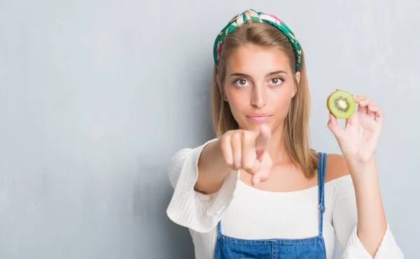 カメラと 指で指している新鮮なキウイを食べてグランジ灰色の壁に美しい若い女性 手に署名 正面からポジティブで自信のジェスチャー — ストック写真