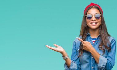 Genç güzel Arap kadın hayrete ve el ile sunulması ve parmak ile işaret ederken kameraya gülümseyen izole arka plan üzerinde güneş gözlüğü takıyor.