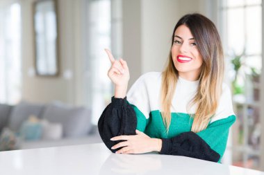 Kameraya bakarak yan el ve parmak ile işaret yüzünde, evde kocaman bir gülümseme ile kış kazak giyen genç güzel kadın.