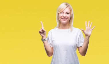 Gösterilen ve parmakları ile işaret izole arka plan üzerinde beyaz t-shirt giyen genç güzel sarışın kadın kendinden emin ve mutlu gülümseyerek süre 6 numara.