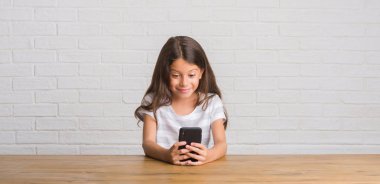 Ayakta ve diş gösterilen kendine güvenen bir gülümsemeyle gülümseyerek mutlu bir yüz ile smartphone kullanarak masada oturan genç İspanyol çocuk