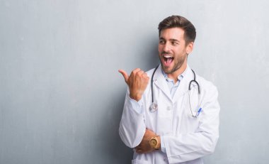 Gri grunge duvara bakarak ve başparmak yanında yukarıyı mutlu yüz ile gülümseyen yakışıklı genç doktor adam.