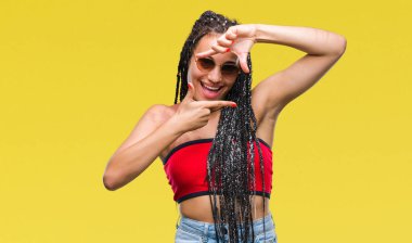 Genç elle yapma çerçeve ve parmakları ile mutlu yüz gülümseyen izole arka plan üzerinde güneş gözlüğü takıyor Doğum işareti ile Afrikalı-Amerikalı saç örgülü. Yaratıcılık ve Fotoğrafçılık kavramı.