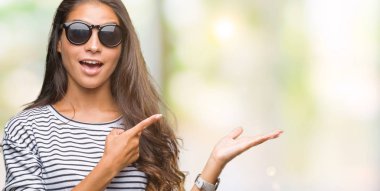 Genç güzel Arap kadın hayrete ve el ile sunulması ve parmak ile işaret ederken kameraya gülümseyen izole arka plan üzerinde güneş gözlüğü takıyor.