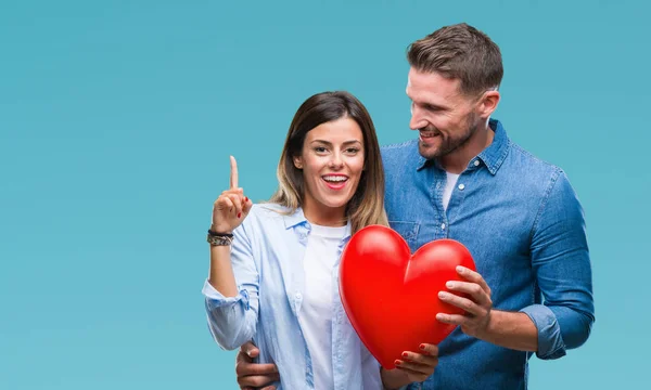 年轻夫妇在爱持有红色心脏在孤立的背景惊讶的想法或问题指向手指与幸福的脸 — 图库照片