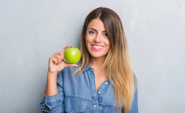 年轻的成年妇女在灰色的粗糙的墙壁上吃着新鲜的绿色苹果与幸福的脸站着 面带微笑 自信的微笑显示牙齿 — 图库照片
