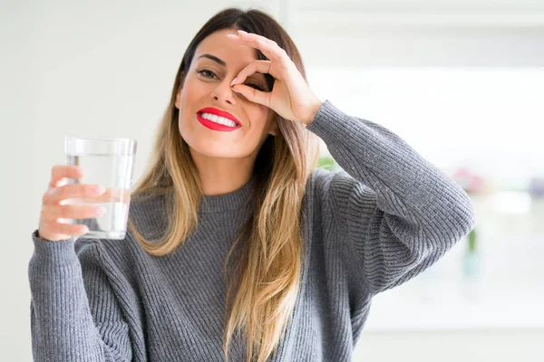 指の間から見て目に手で サインをして笑っている幸せそうな顔と家庭での水のガラスを飲む若い美しい女性 — ストック写真