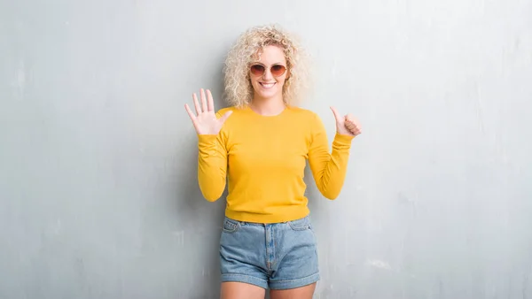 Young blonde woman with curly hair over grunge grey background showing and pointing up with fingers number six while smiling confident and happy.