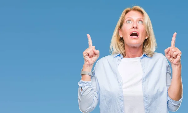 Middle age blonde woman over isolated background amazed and surprised looking up and pointing with fingers and raised arms.