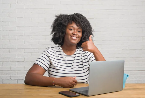 Young african american woman sitting on the table using computer laptop happy with big smile doing ok sign, thumb up with fingers, excellent sign