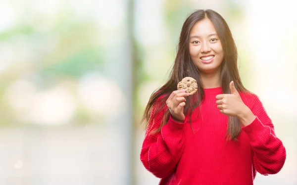 笑顔の サイン 優秀な兆候を親指をやって幸せな孤立の背景の上のチョコレート チップ クッキーを食べる若いアジア女性 — ストック写真